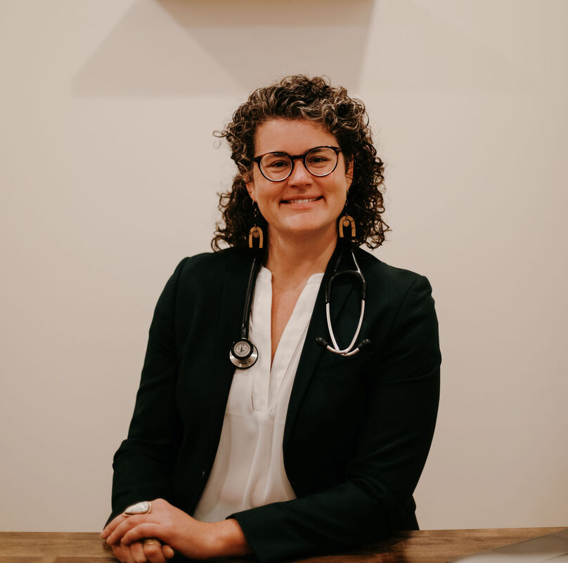Dr. Barrett, a white cis-female with brown curly hair, smiling. She wears dark, heavy glasses and a turquoise shirt with a black blazer.