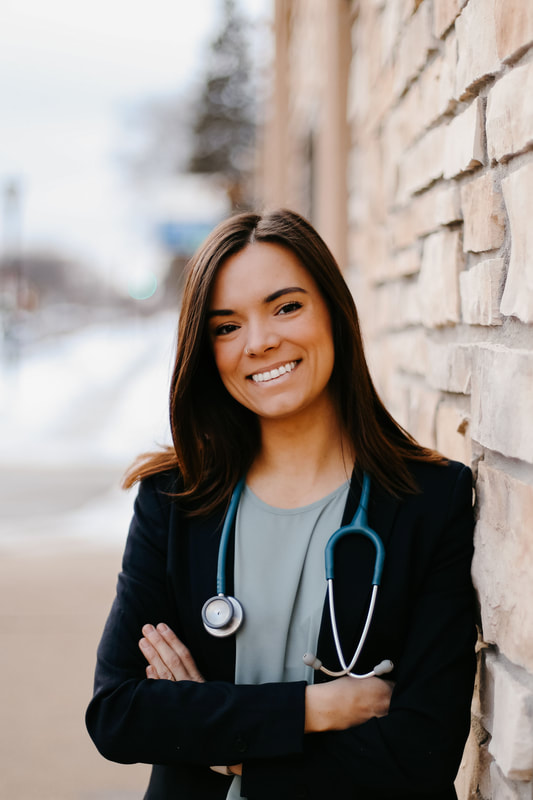 Dr. Aidanne, a cis-female naturopathic doctor with brown hair wearing a blue dress and standing in front of green foliage