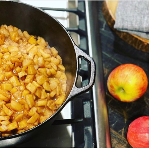 Cast iron skillet on a stove with cut apples cooking inside and 2 whole red apples on the countertop next to the stove