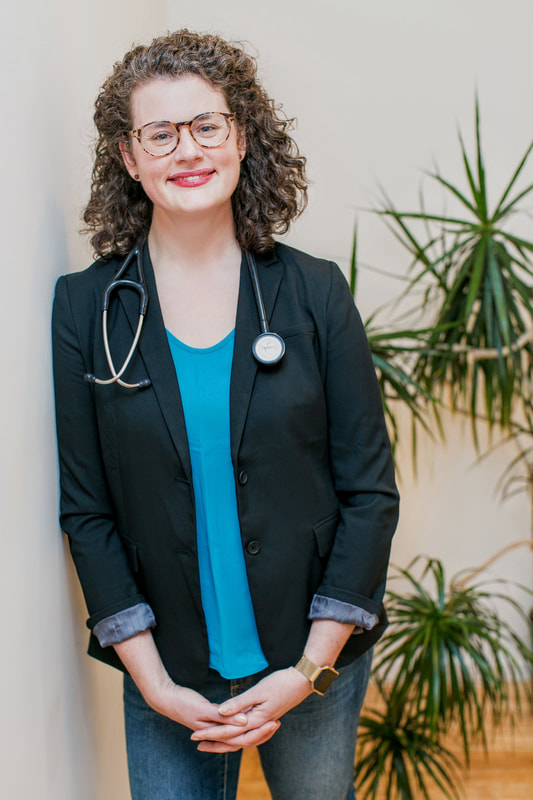 Dr. Barrett, a white cis-female with brown curly hair, smiling. She wears dark, heavy glasses and a turquoise shirt with a black blazer.